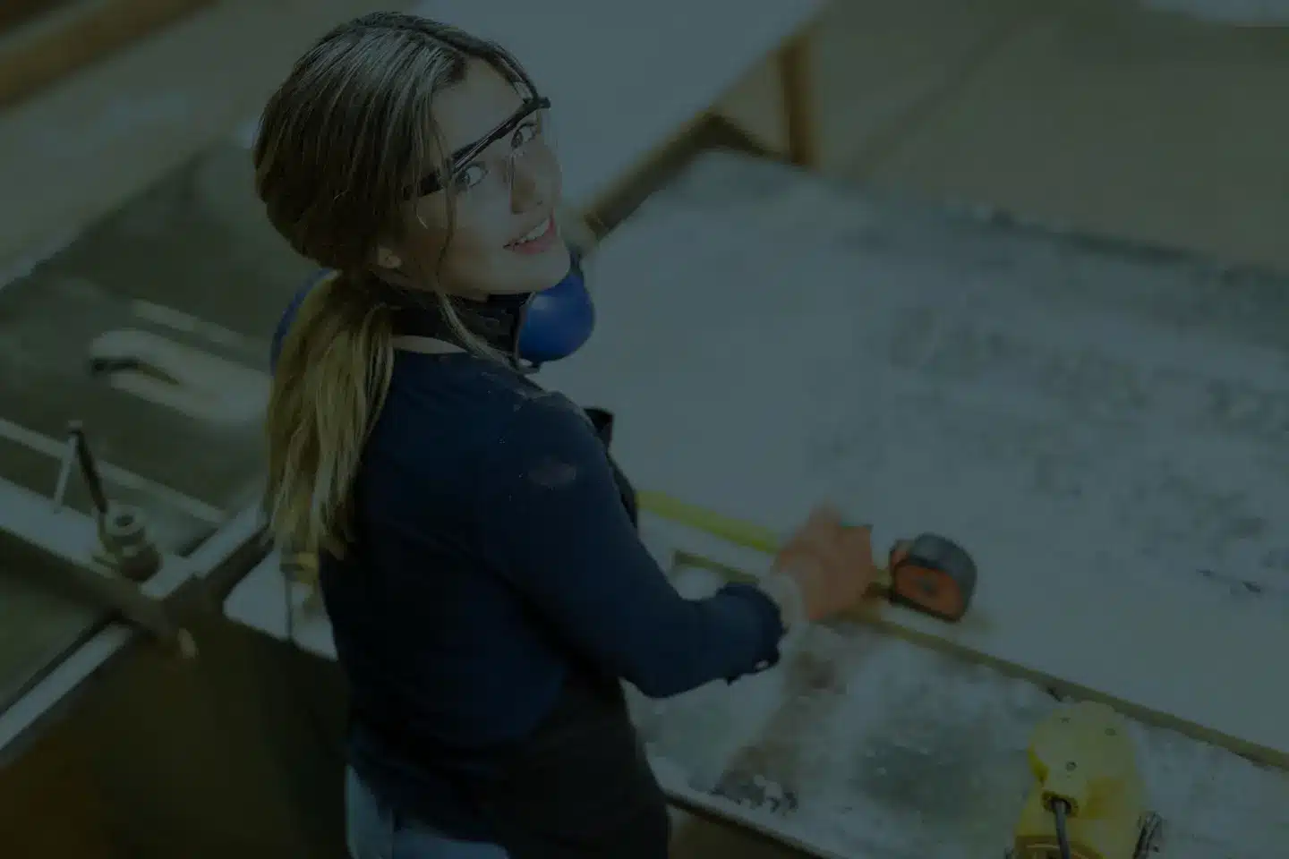 Junge Handwerkerin mit Schutzbrille misst Holzbrett in Werkstatt – freundliches Motiv für Recruiting und Außenwerbung Koblenz